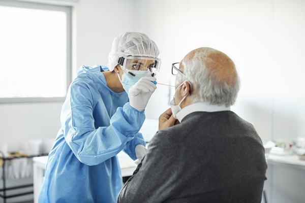 In the color photo, a person wearing blue places a cotton swab in the mouth of a seated elderly man.