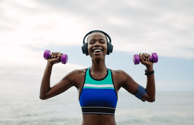 Happy woman wearing earphones exercising with dumbbells outdoors.  Female fitness wear headphones workout done at the beach in the morning.  (Photo: Getty Images/iStockphoto)
