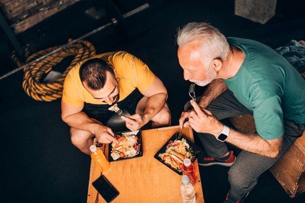 In the color image, two men occupy the center of the image.  They eat from black plates and wear yellow and green blouses.